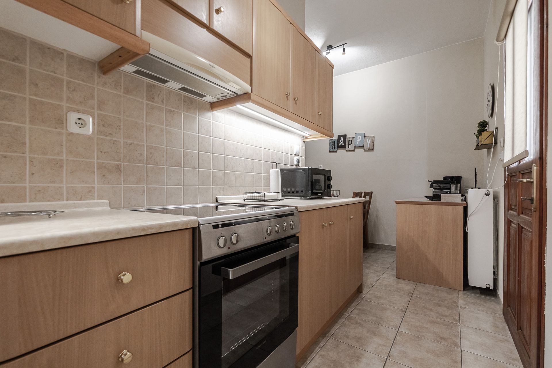 Narrow beige kitchen with wood cabinets, tiled floor, stove, countertop, and small appliances