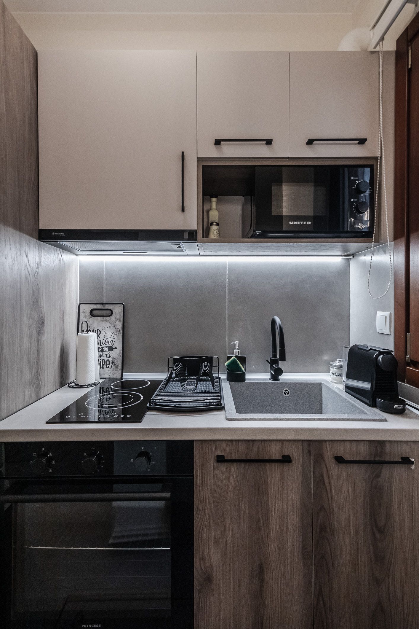 Compact modern kitchen with wood cabinets, stainless sink, stove, and microwave under under-cabinet lighting.
