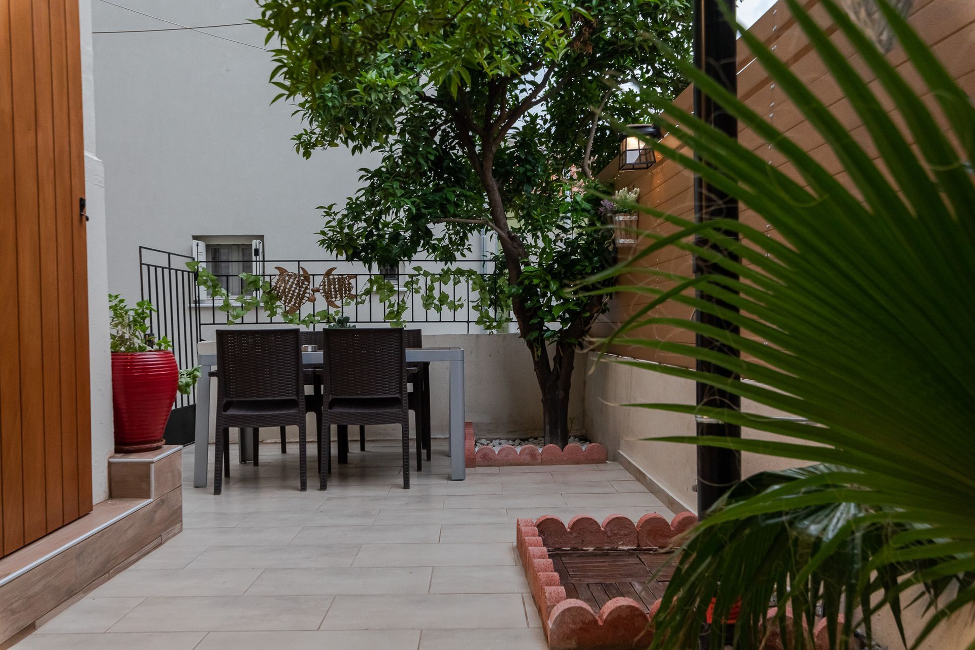 Patio with table and chairs, potted plants, white walls, and a leafy tree in a courtyard.