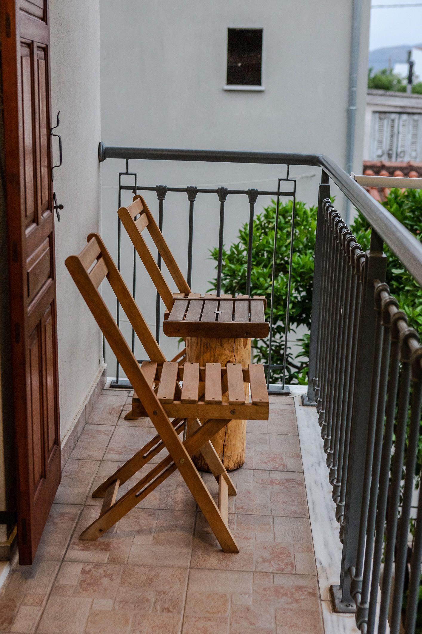 Small balcony with wooden folding chairs and table beside a railing and potted greenery.
