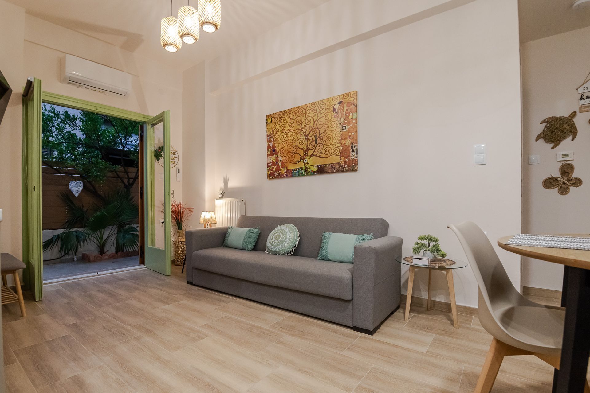 Bright living room with gray sofa, wall art, open green doors, and dining area with chairs.