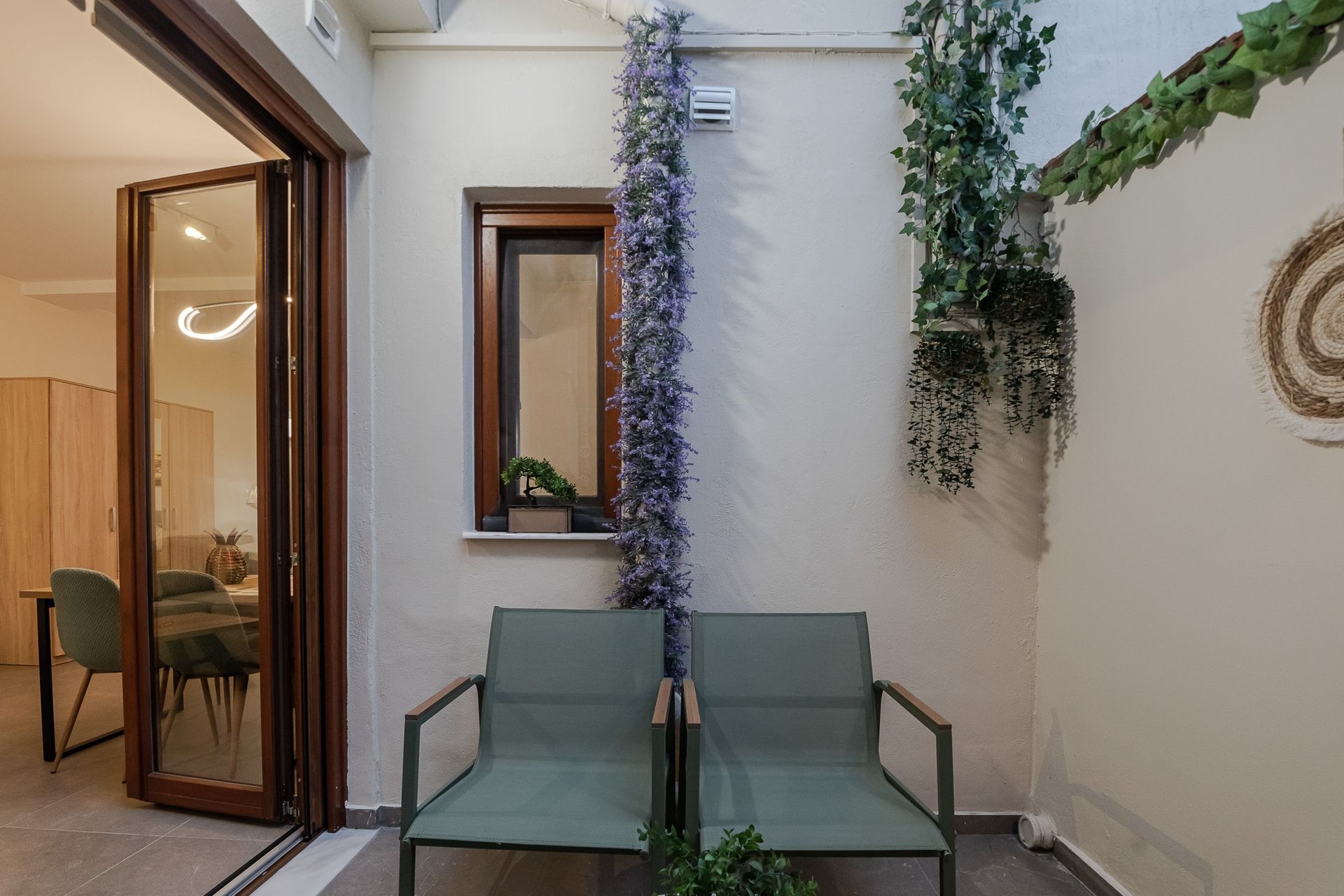 Patio seating area with two green chairs, potted plants, and an open glass door leading inside