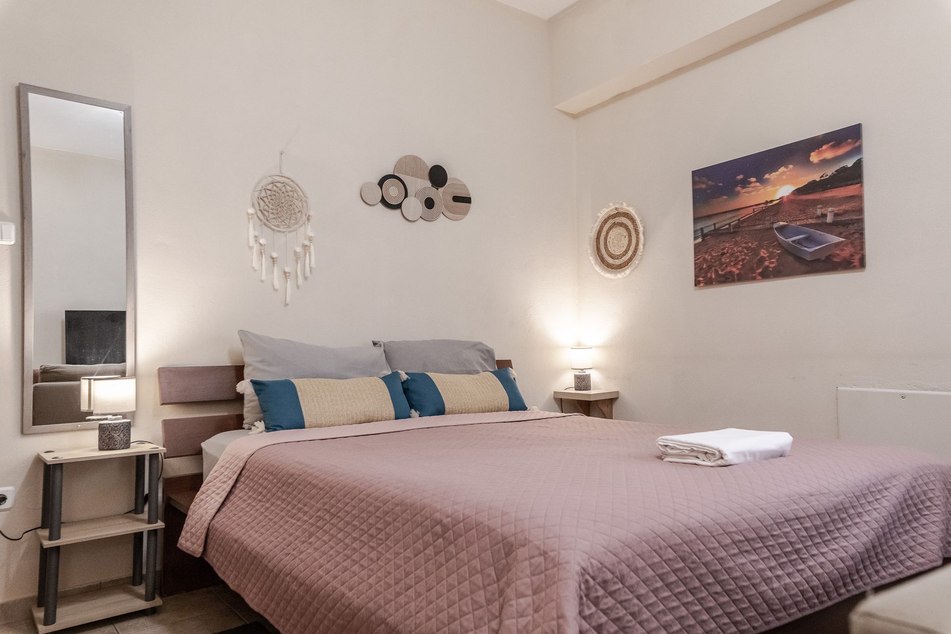 Cozy bedroom with a bed, pink quilt, blue pillows, bedside tables, mirror, and wall decor