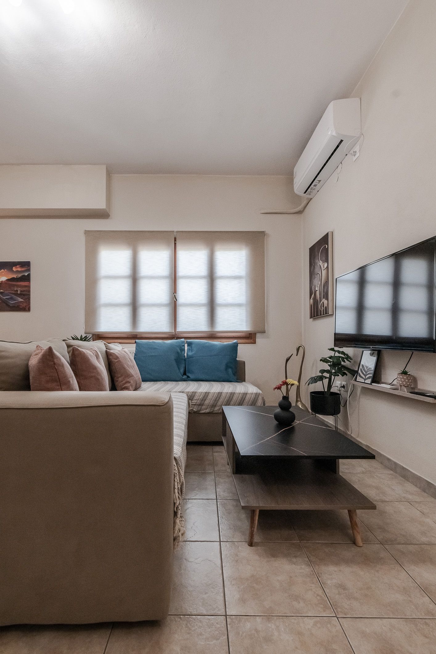Modern living room with beige sectional, blue cushions, TV, coffee table, window, and wall-mounted AC unit