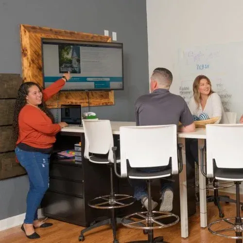 Woman points at a wall-mounted screen during a small office meeting with two seated people.
