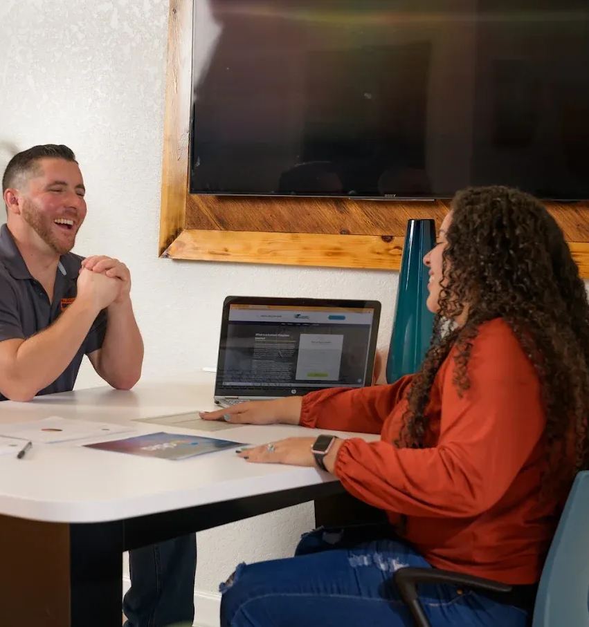 Two people in a meeting at a table, talking beside an open laptop in a bright room.