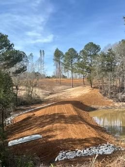 Curving dirt road beside a pond, with piles of gravel and tall pine trees under a blue sky.