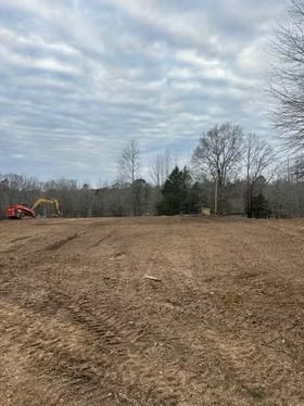 Cleared dirt field with excavators at left under cloudy sky, bordered by leafless trees