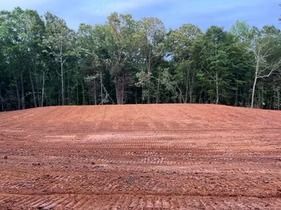 Freshly graded red dirt lot bordered by dense green trees under a blue sky