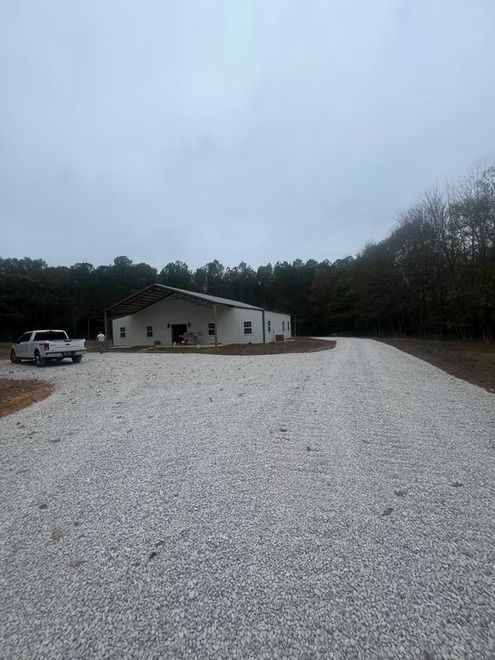 Gravel driveway leading to a white building with parked vehicles under an overcast sky, surrounded by trees.