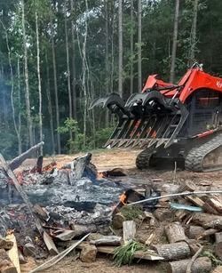 Excavator beside a smoldering debris pile in a forest clearing, with tall trees in the background