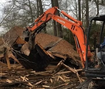 Orange excavator demolishing a collapsed wooden building amid rubble and debris outdoors