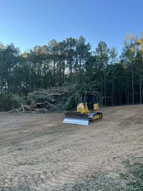 Yellow bulldozer clearing a dirt lot beside a wooded area under a blue sky