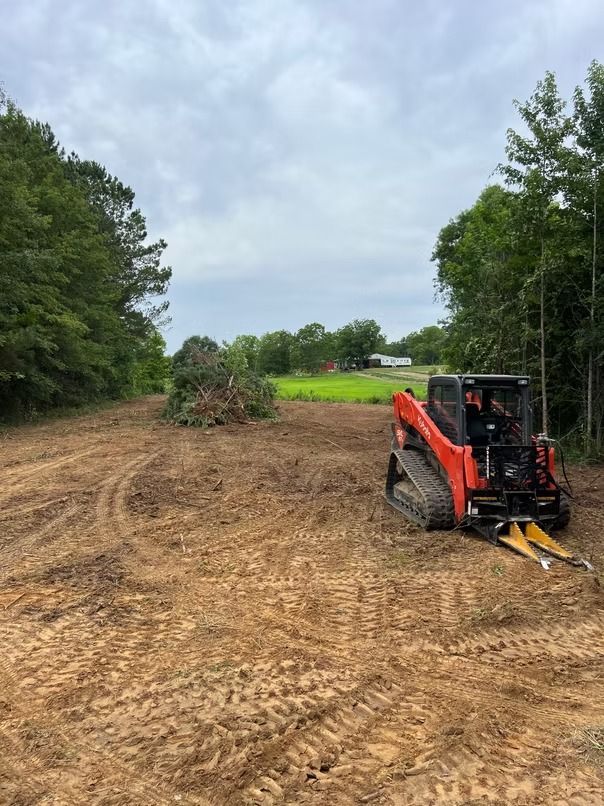 Orange skid steer grading a dirt lot beside trees under a cloudy sky