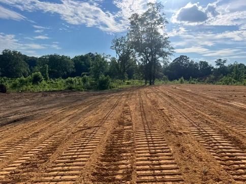 Freshly graded dirt field with tire tracks, bordered by trees under a partly cloudy sky