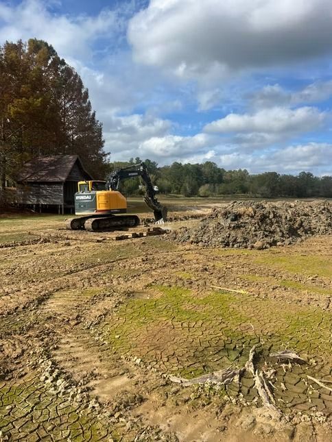 Yellow excavator clearing a muddy field near a barn under a cloudy sky