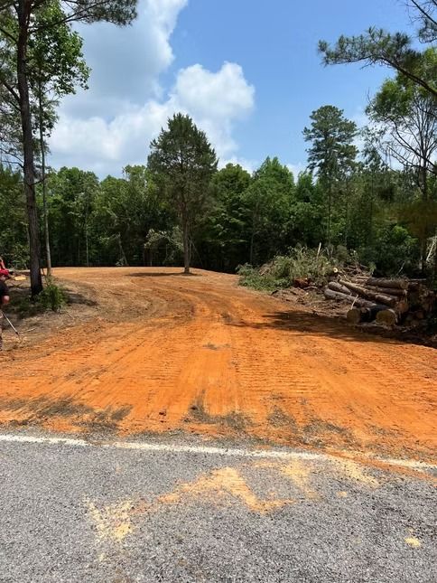 Orange dirt road leading into a wooded area, with piled branches on the right and a paved road in front