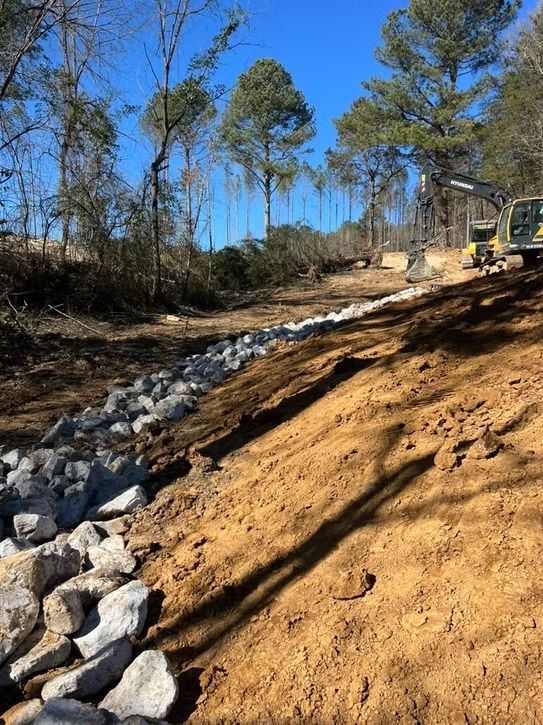 Construction site with excavator shaping a dirt slope beside a rocky drainage ditch and pine trees