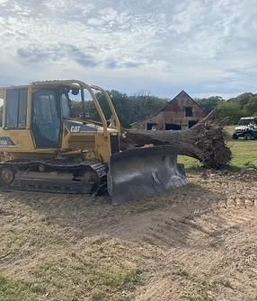 Yellow Caterpillar bulldozer moving a fallen tree trunk near a barn.
