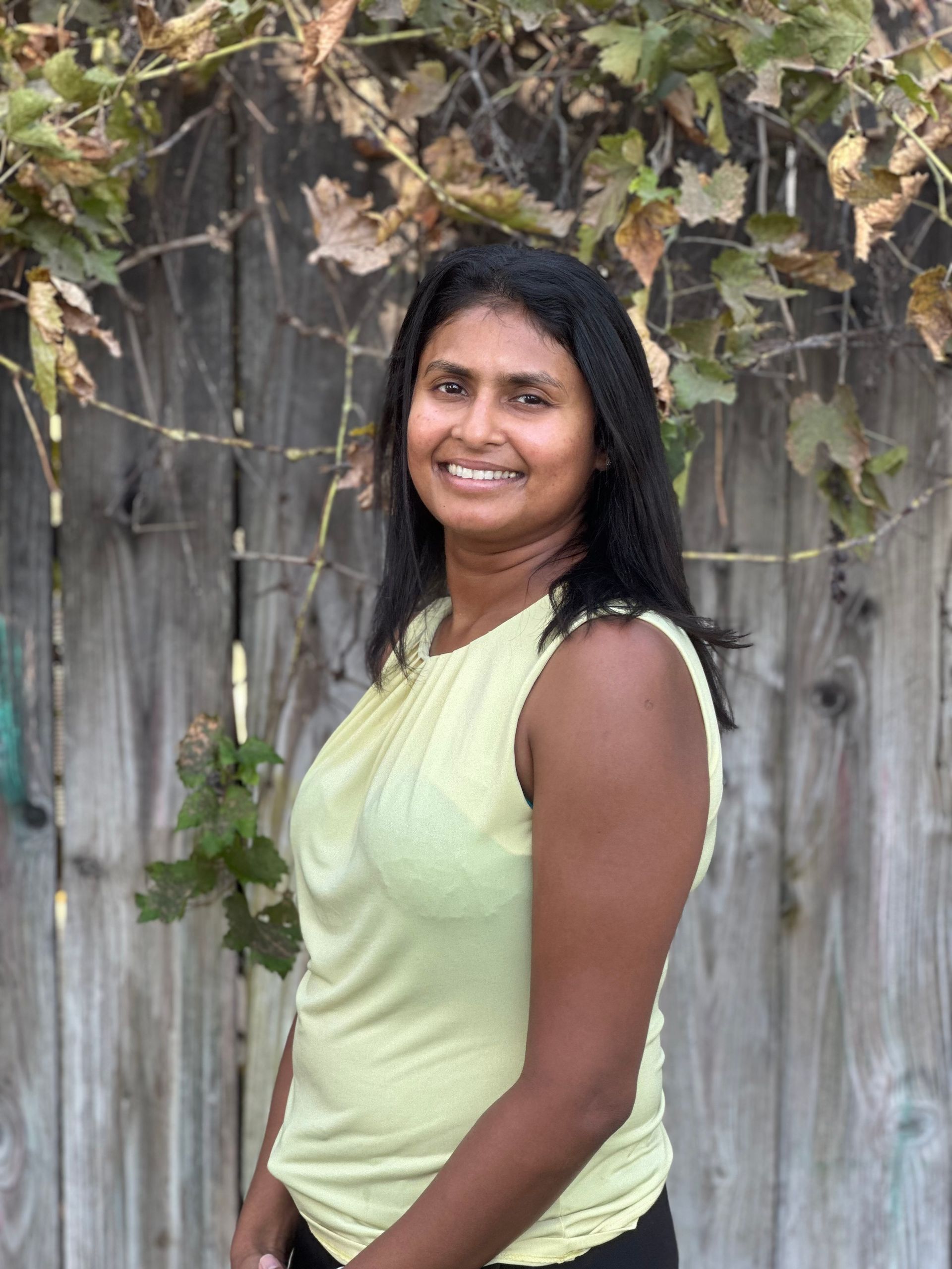 A woman in a yellow tank top is smiling in front of a wooden fence.