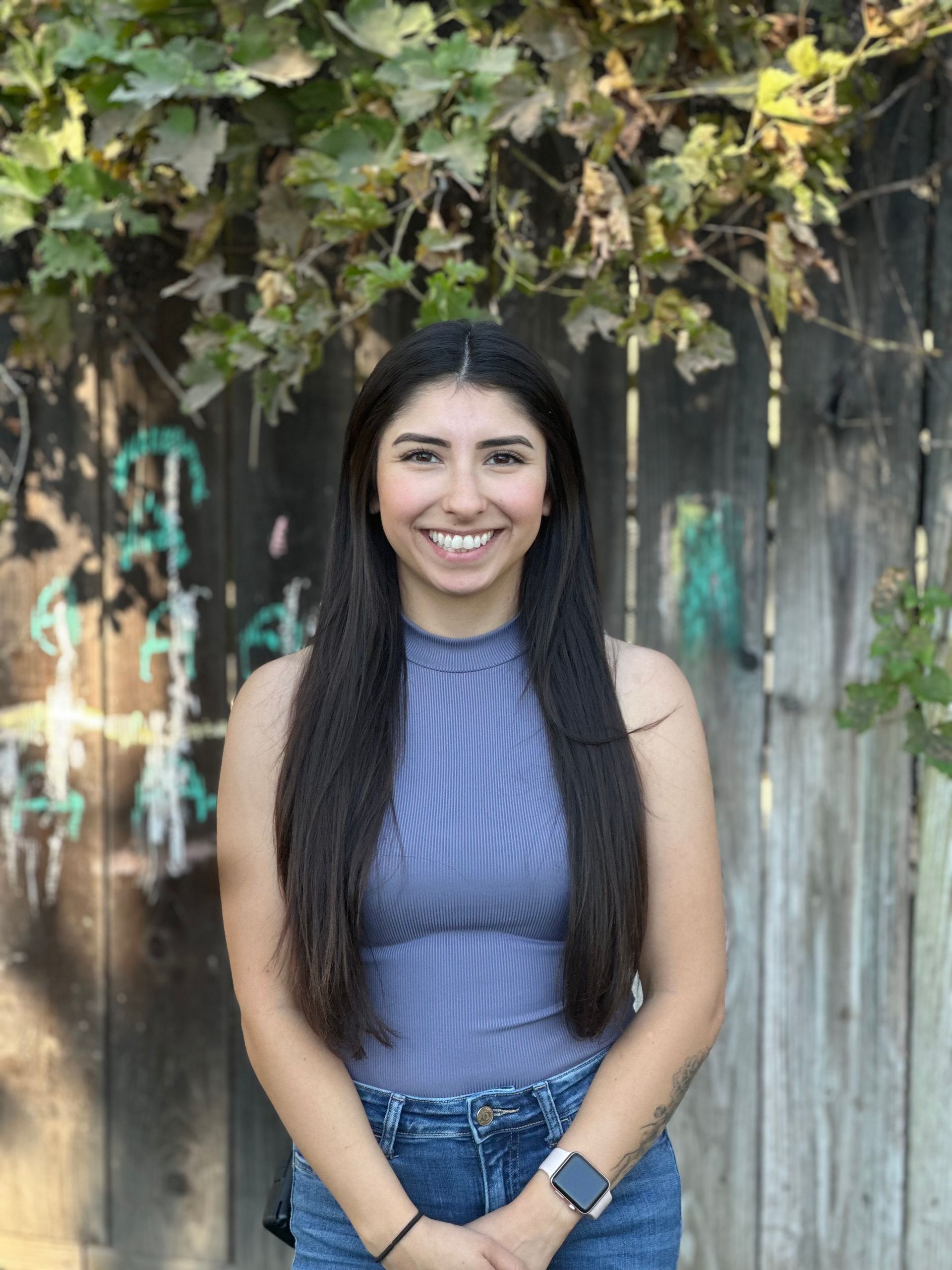 A woman with long hair is standing in front of a wooden fence and smiling.