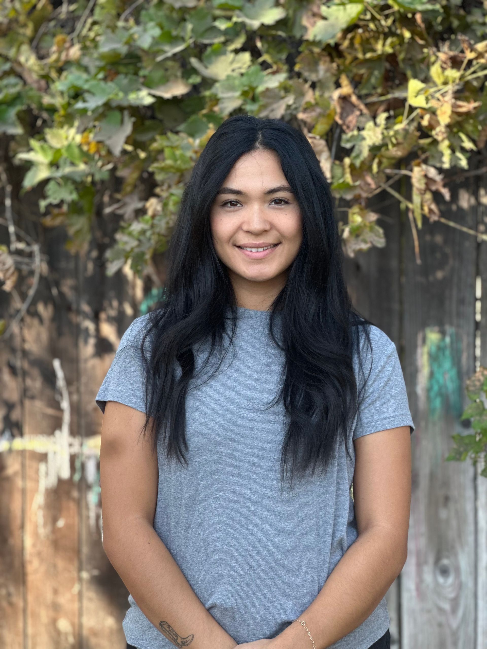 A woman in a grey shirt is standing in front of a wooden fence.