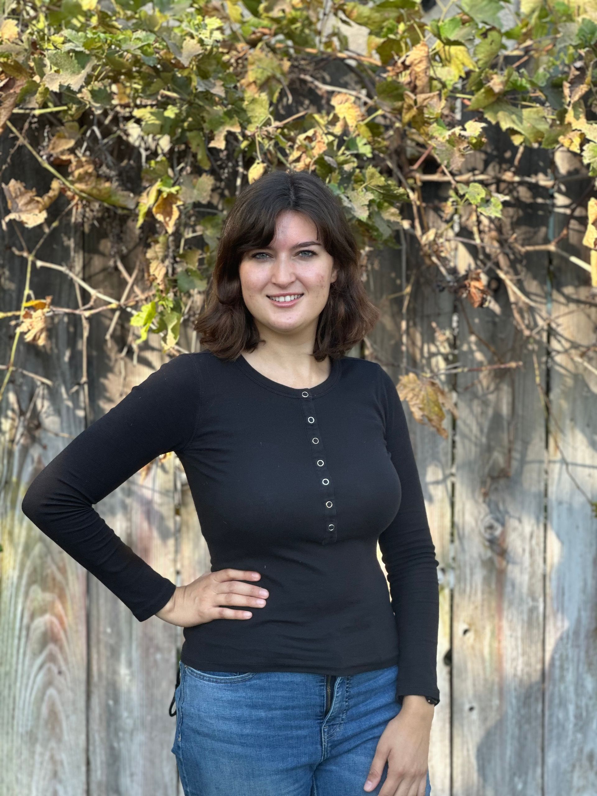 A woman in a black shirt and blue jeans is standing in front of a wooden fence.