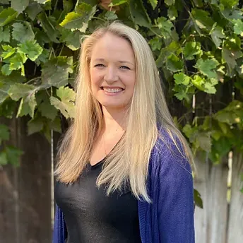 A woman in a black shirt and blue cardigan is smiling in front of a tree.