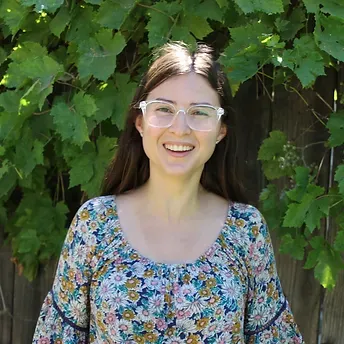 A woman wearing glasses and a floral shirt is smiling in front of a vine.