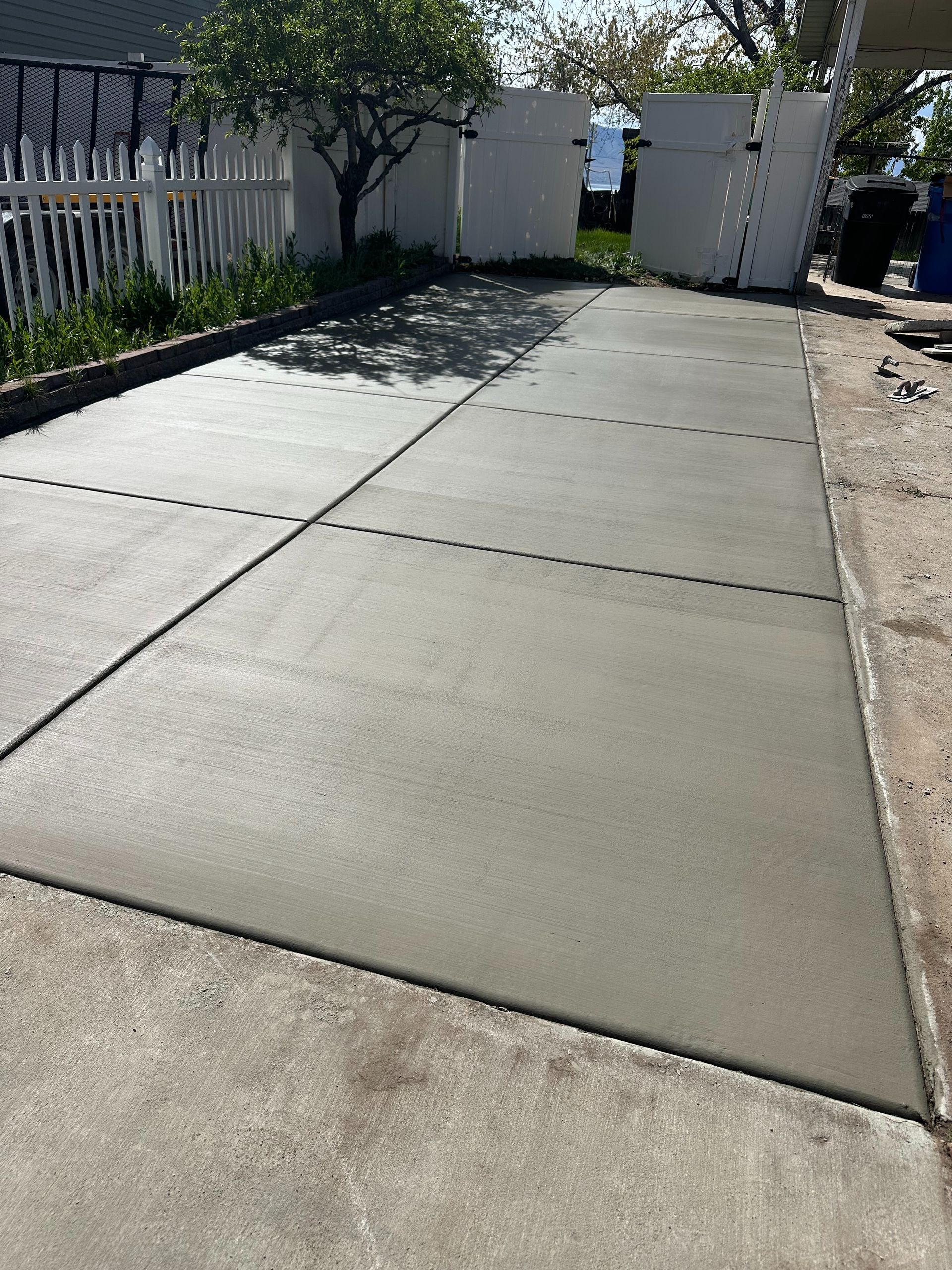 Concrete driveway leading to a white gate beside a fence and trees in bright sunlight
