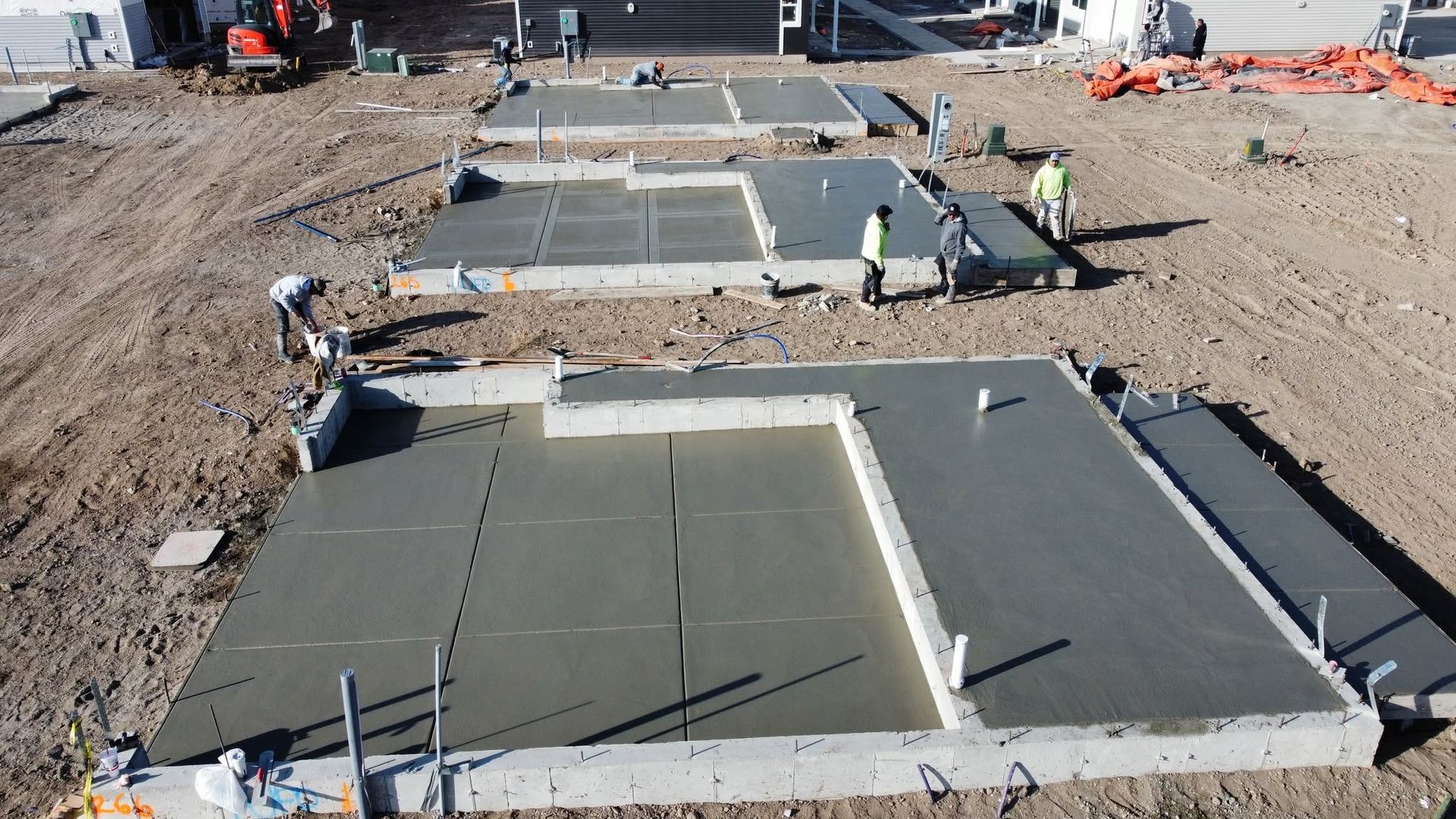 Concrete slabs being poured at a construction site, with workers smoothing wet concrete and dirt around the foundation.