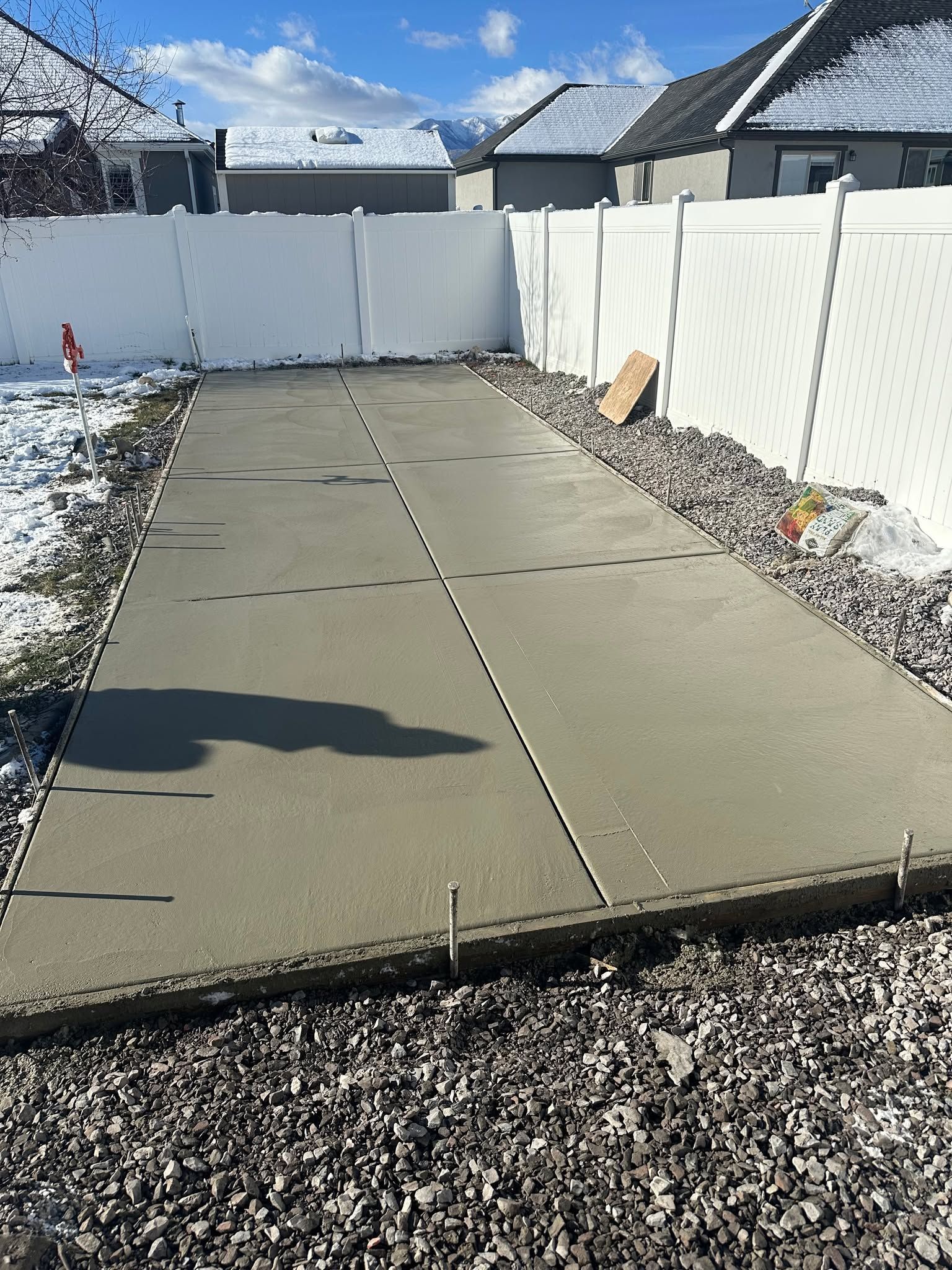 Concrete driveway or patio beside a white fence with snow on the ground under a bright blue sky.