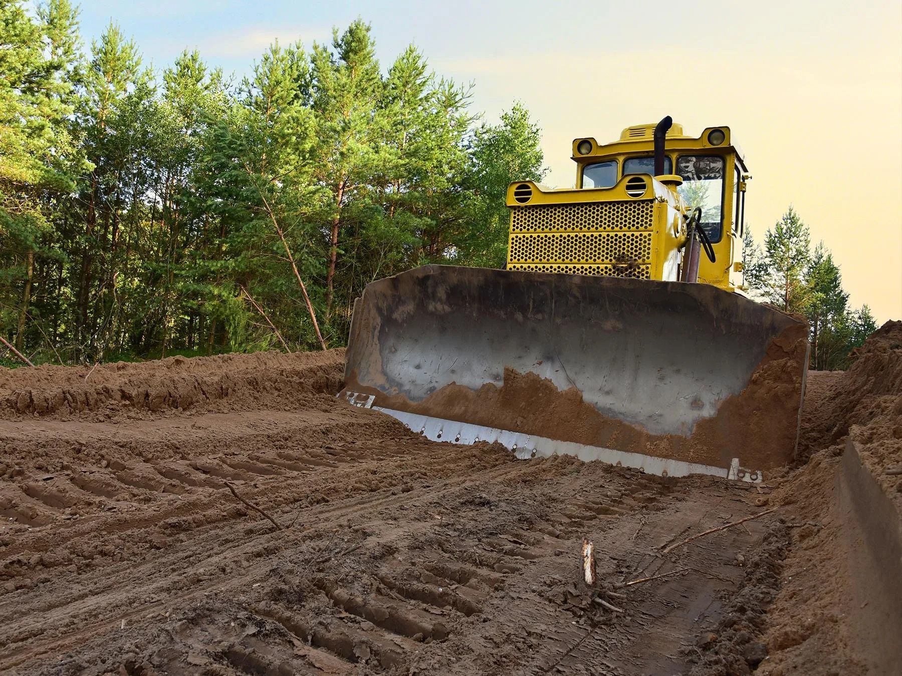 Yellow bulldozer grading a dirt road beside trees under a clear sky