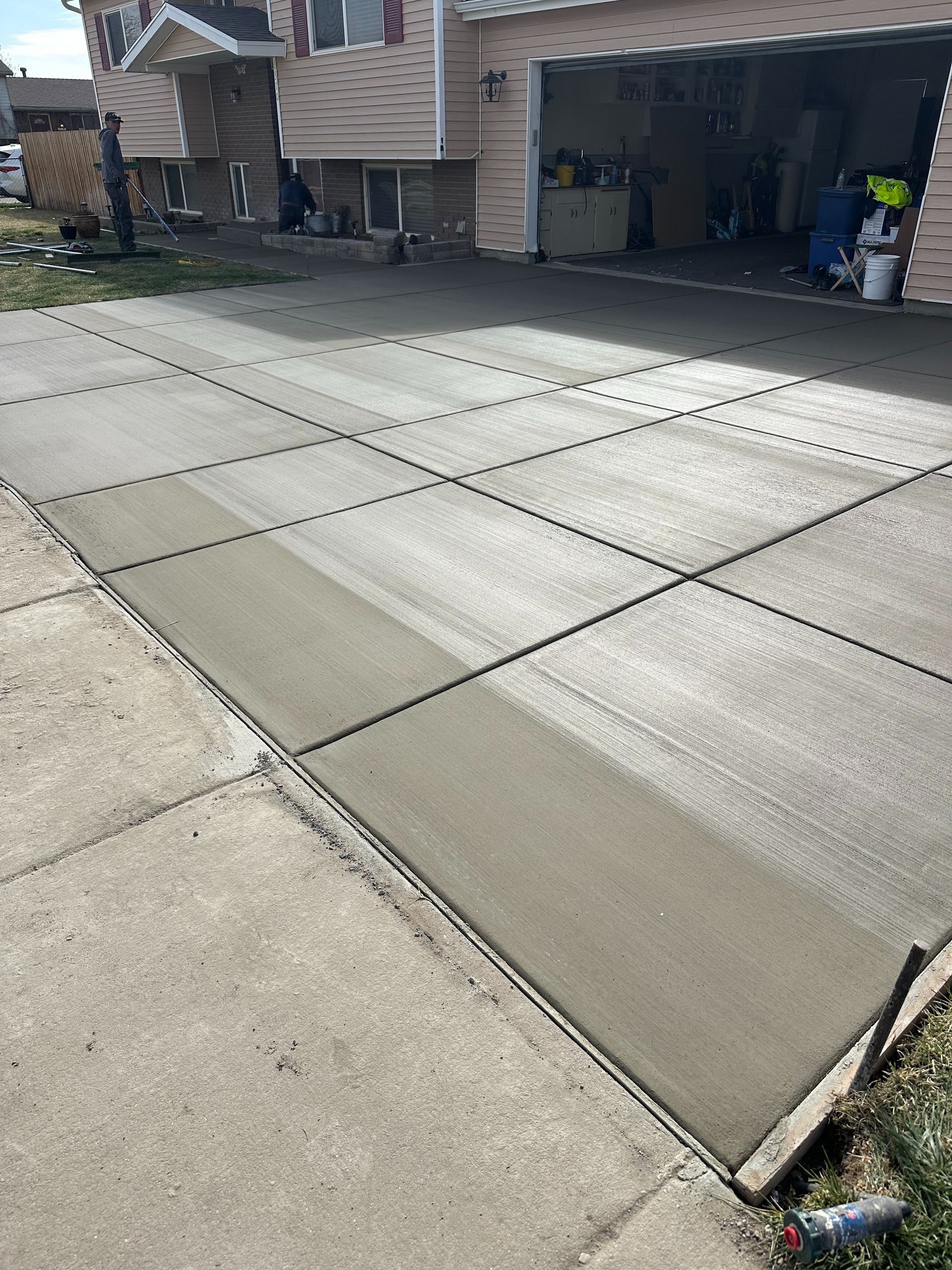 Freshly poured concrete driveway in front of a garage, with workers visible near the open bay.