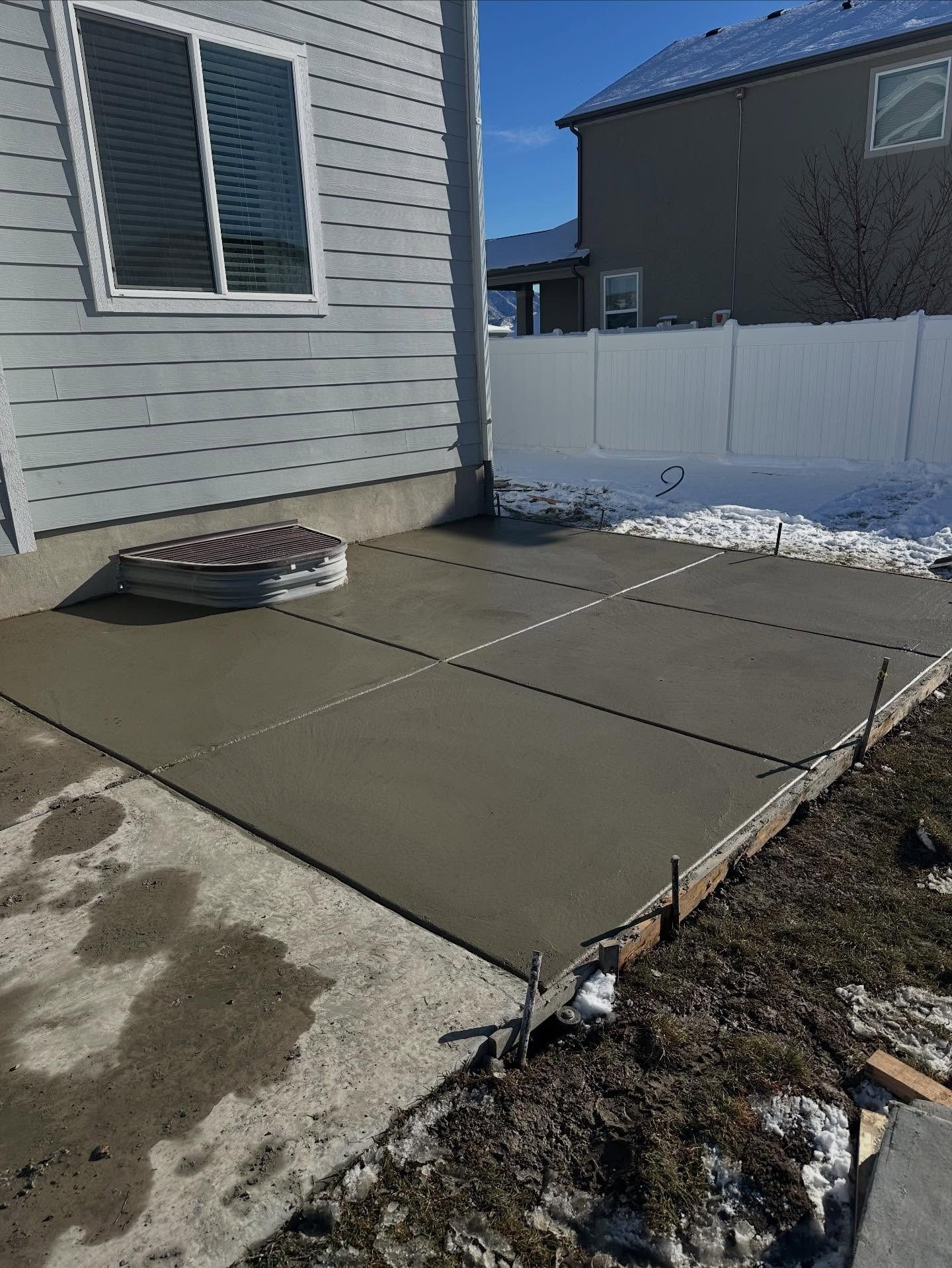 Concrete patio slabs beside a house and white fence, with patches of snow on the ground.