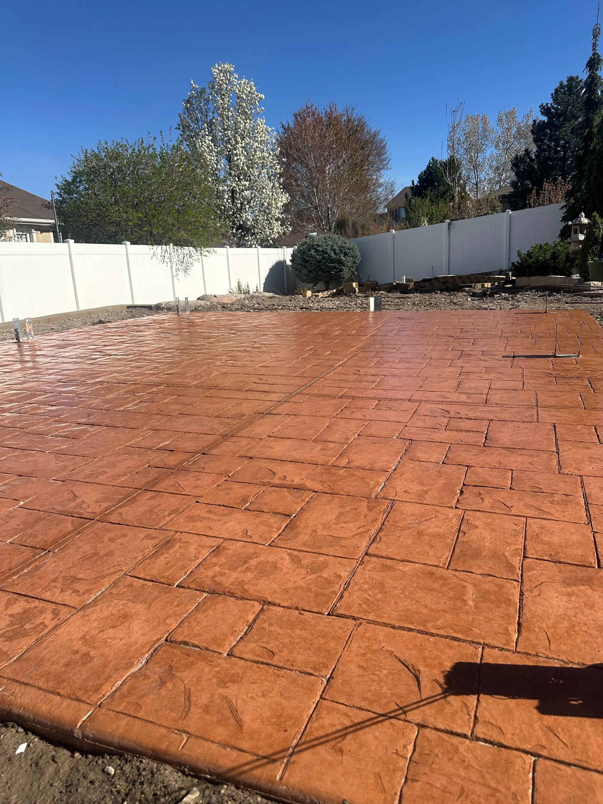 Red brick patio in a fenced backyard under a clear blue sky, with trees in the background.