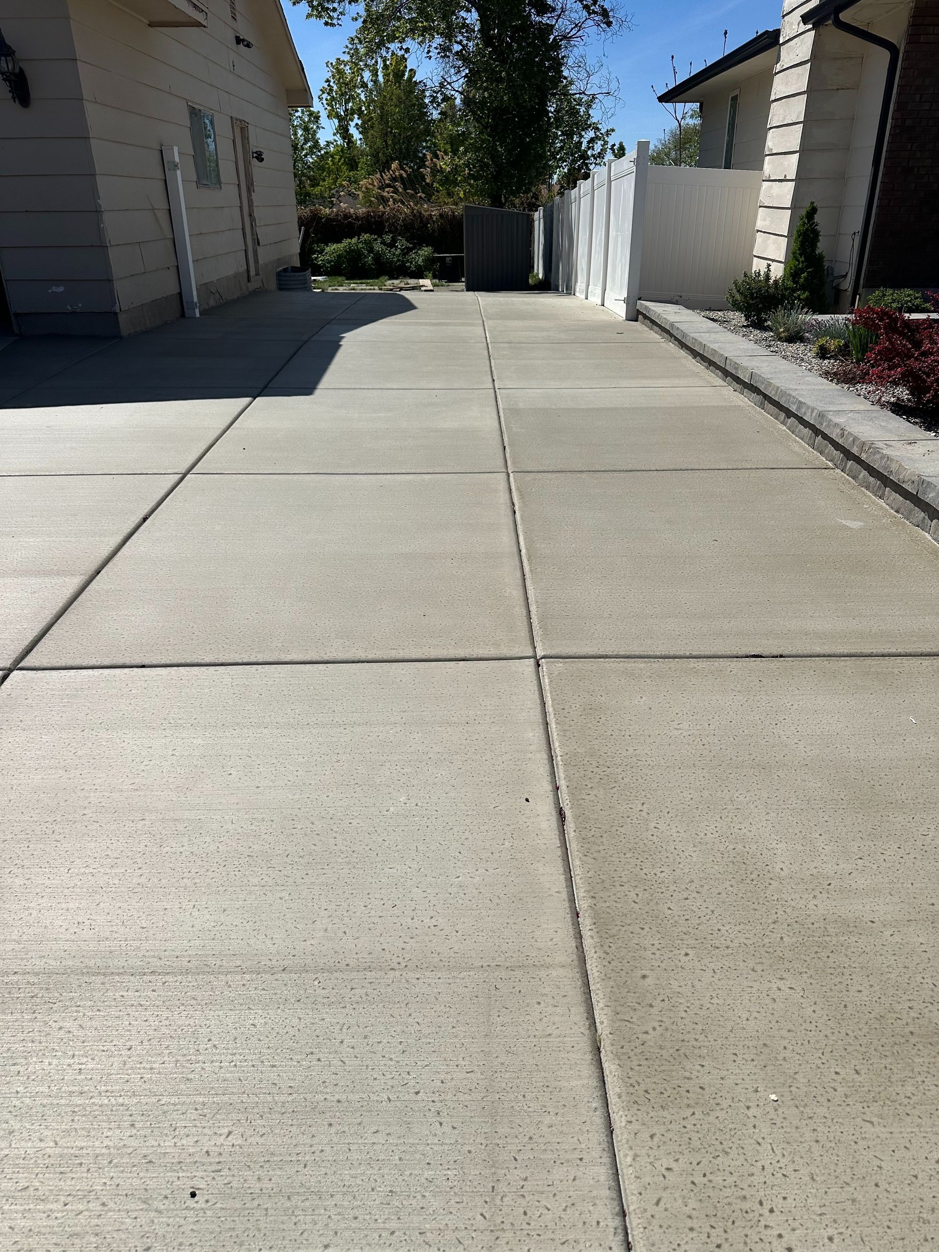 Sunlit concrete walkway between residential buildings, leading to a shaded gate and trees.