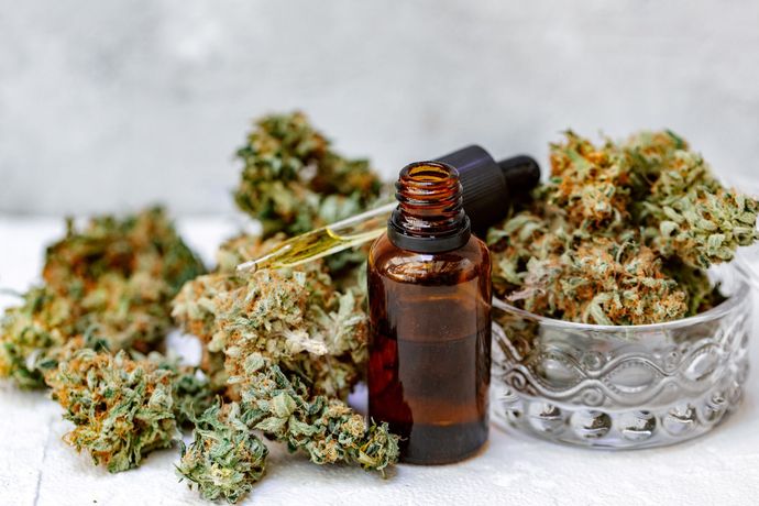 CBD oil dropper bottle beside cannabis buds in a glass bowl on a white surface