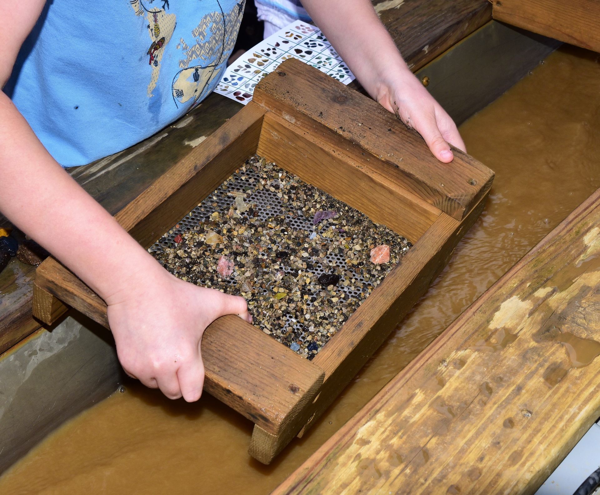 Hands holding a wooden pan with gravel, possibly panning for gems in a water trough.