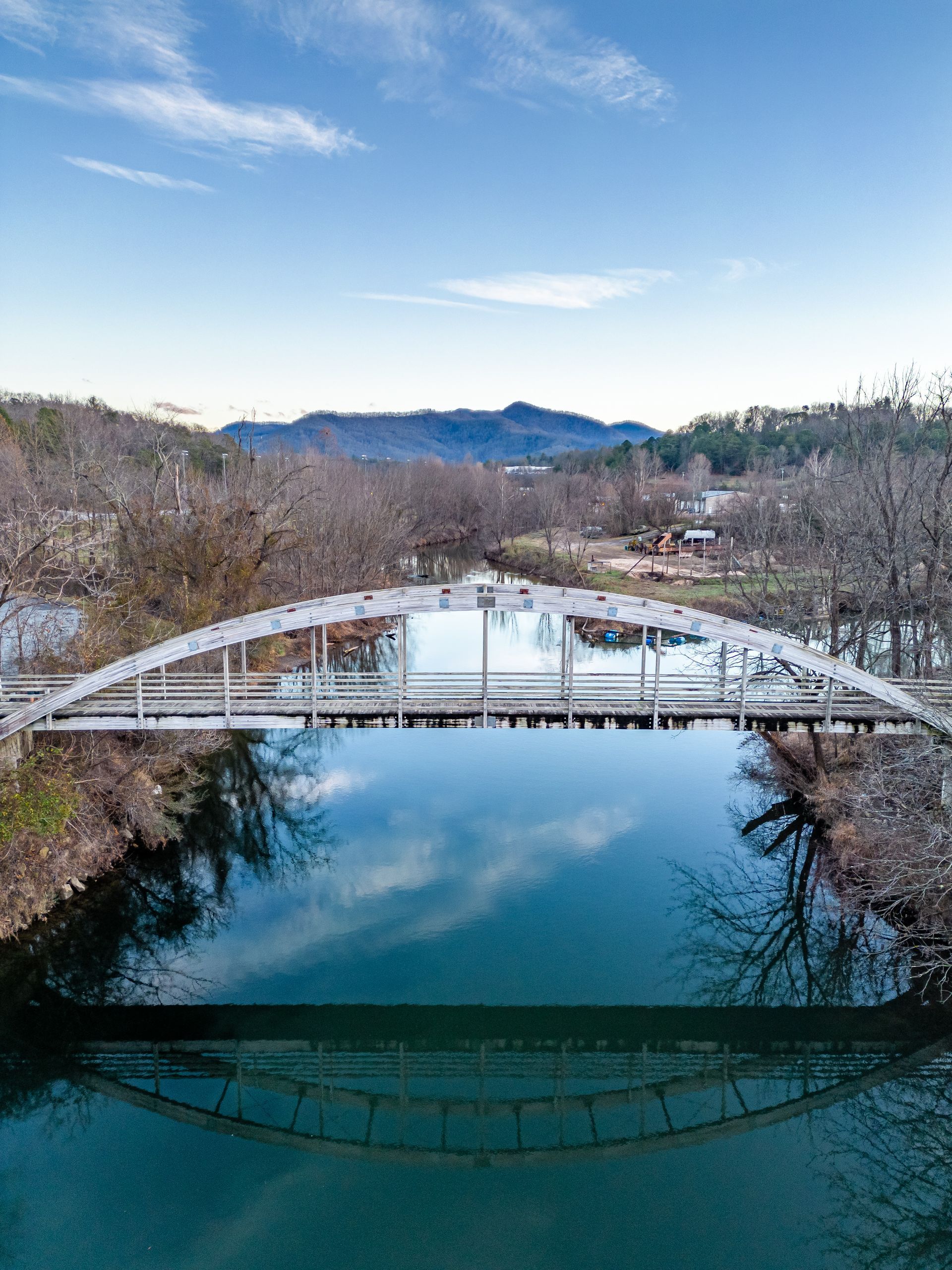 A white arched bridge reflects in a calm blue river, with trees and mountains under a clear sky.