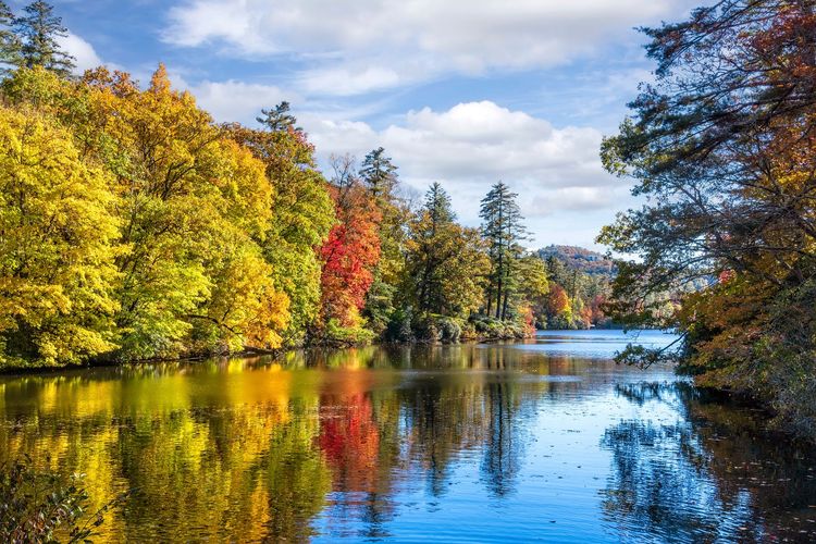 Autumn trees with vibrant yellow, orange, and red leaves reflected on a calm lake under a blue sky.