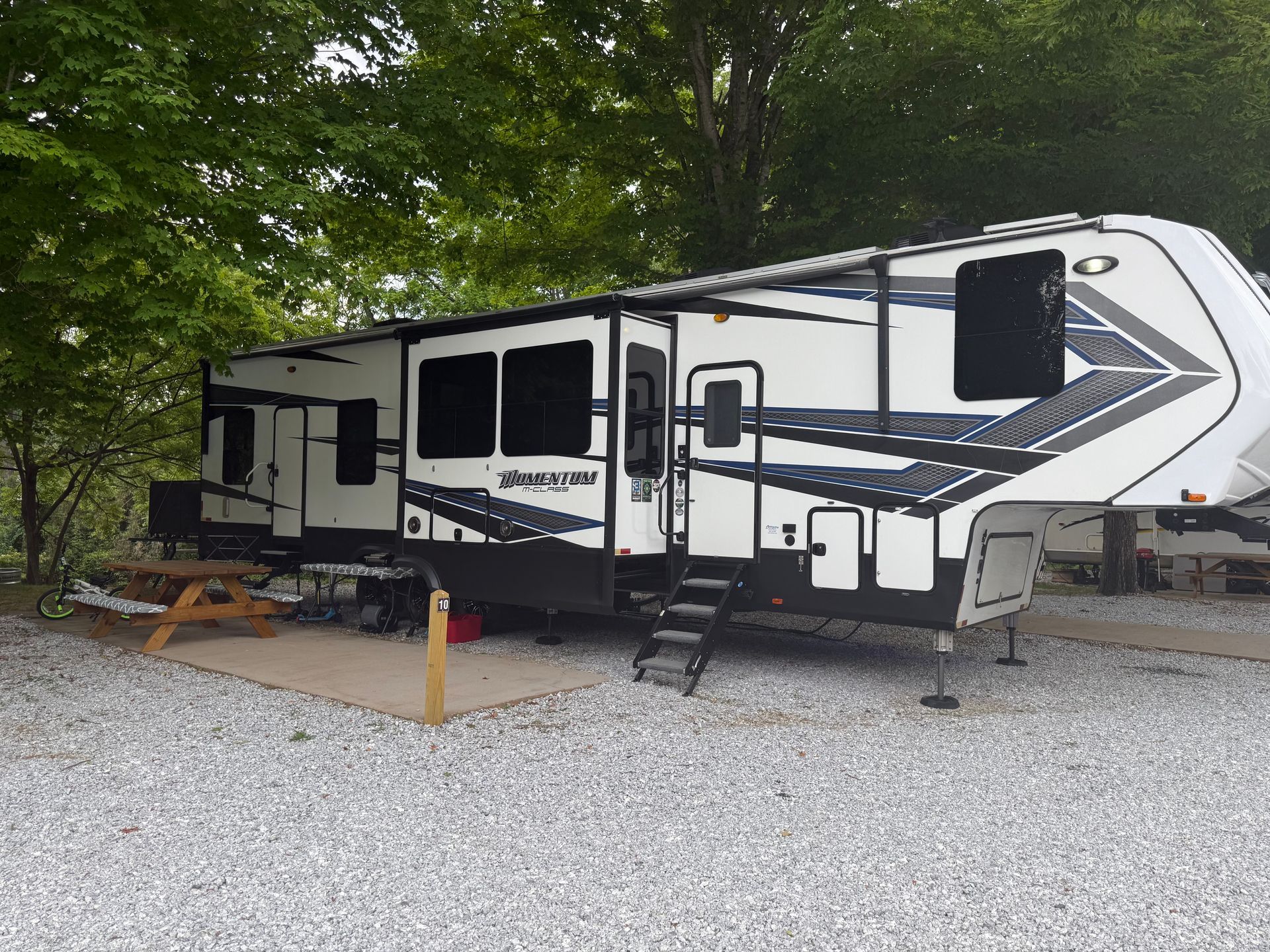 Large RV parked on gravel, with picnic table and steps. White and black design, trees in background.
