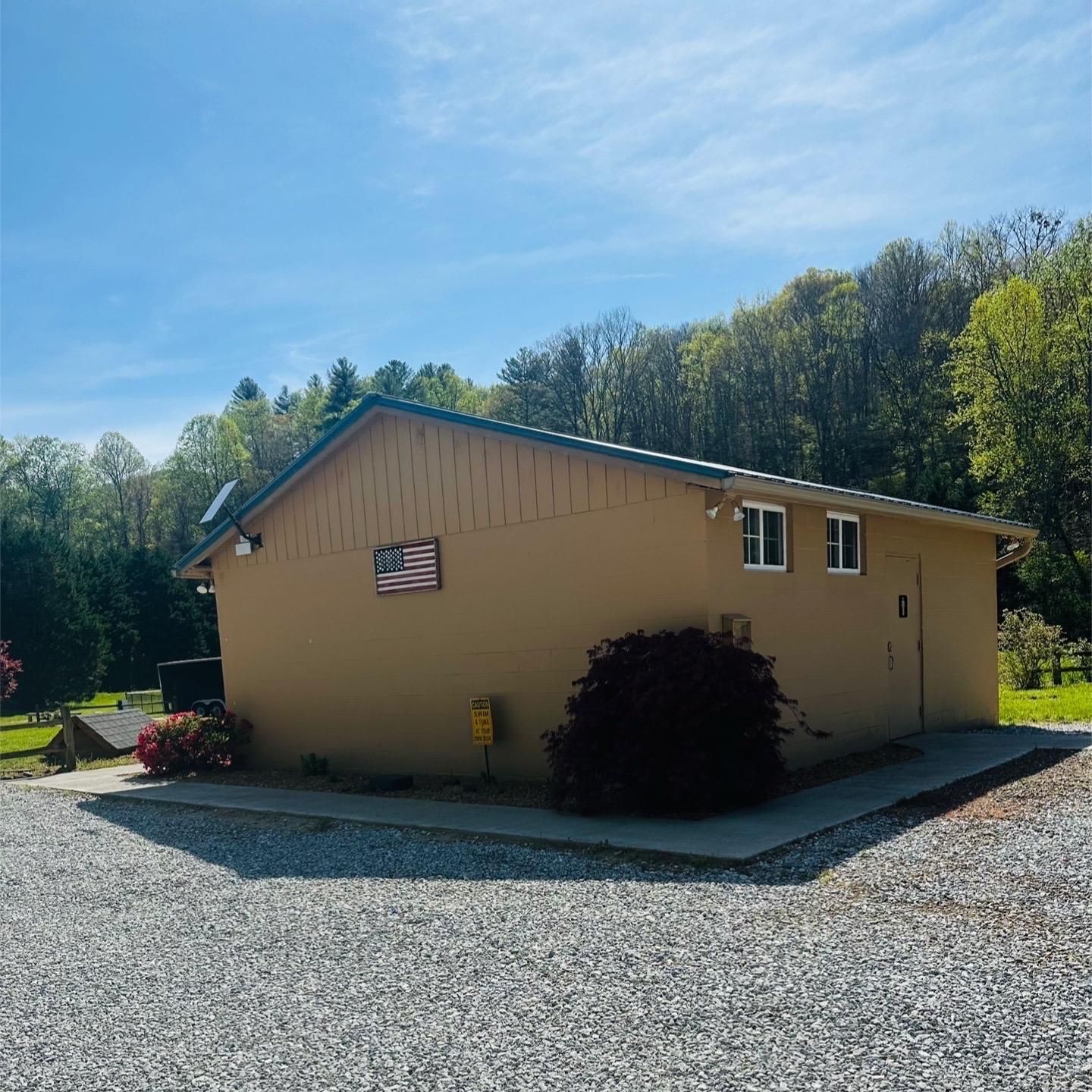Tan building with American flag, two windows, and gravel driveway. Trees in background.