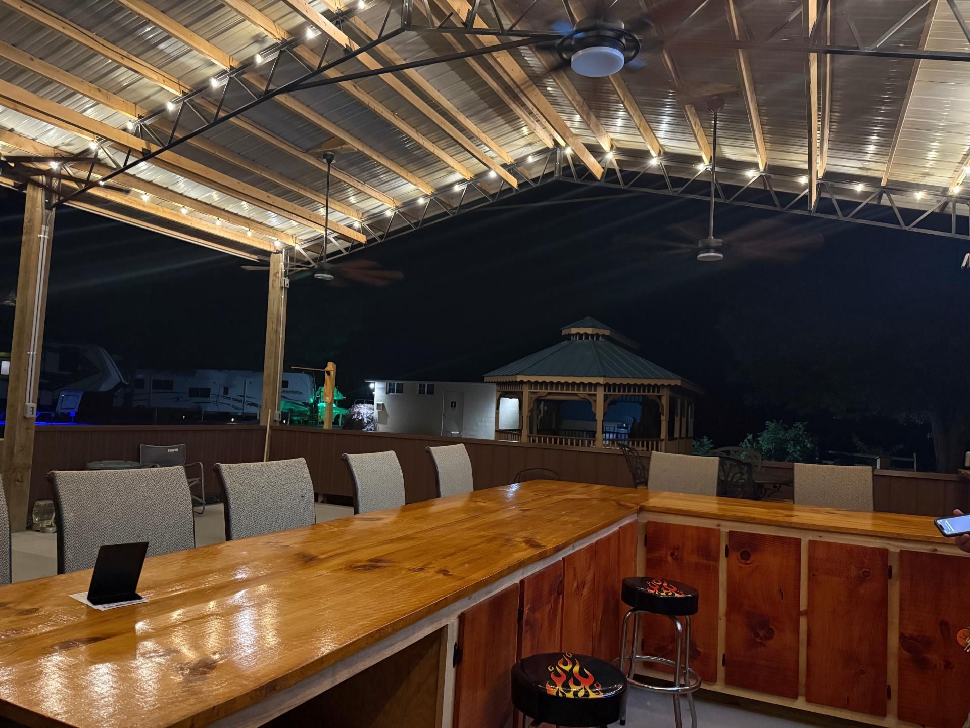 Outdoor bar at night with string lights, wooden counters, bar stools, and a gazebo in the background.