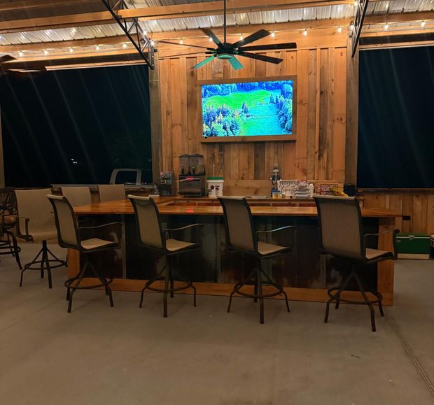 Outdoor bar with seating, TV, and ceiling fan against a wooden wall.
