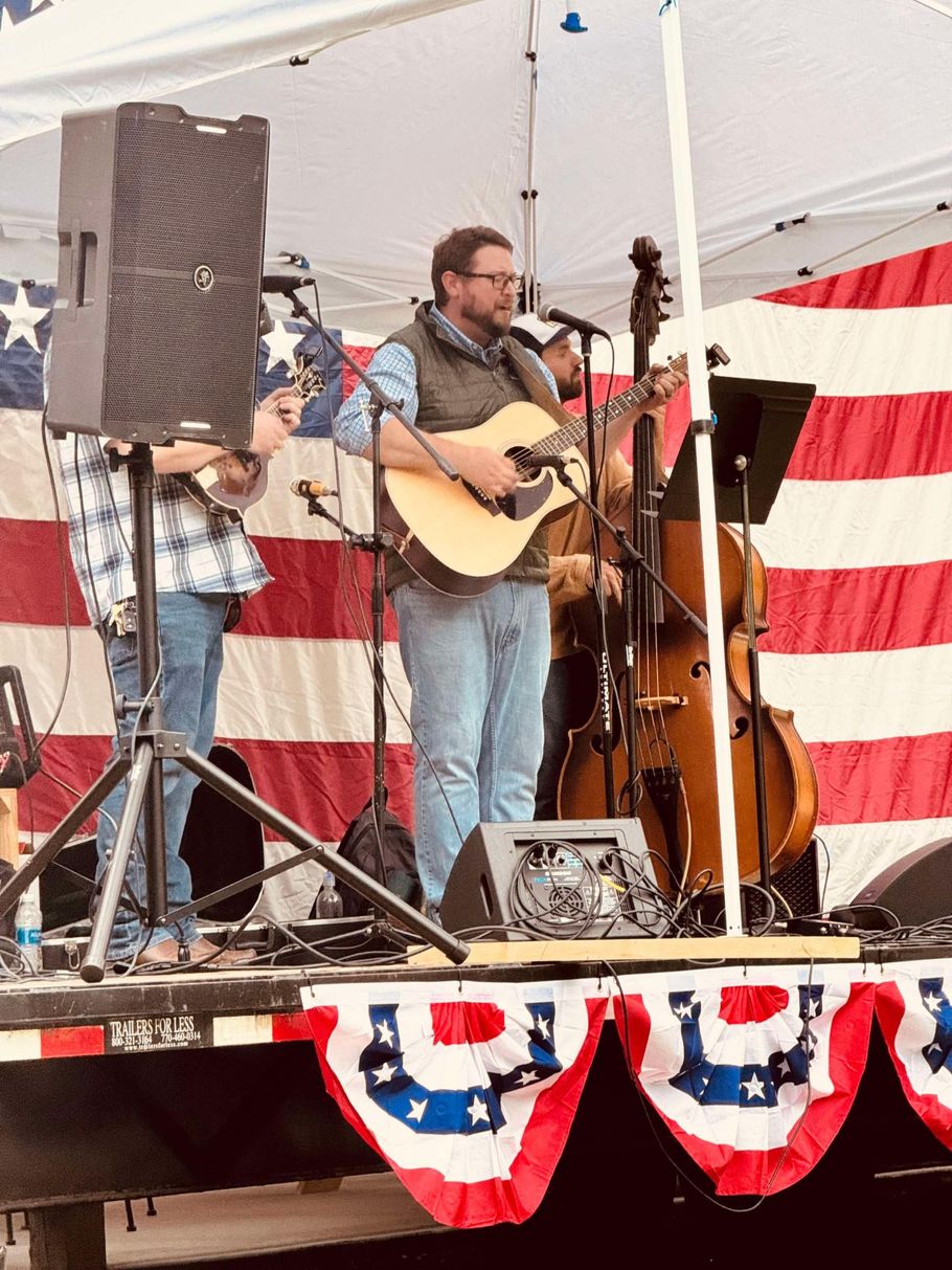 A band performs on stage. Man sings and plays guitar, backed by upright bass and other musicians. American flag backdrop.