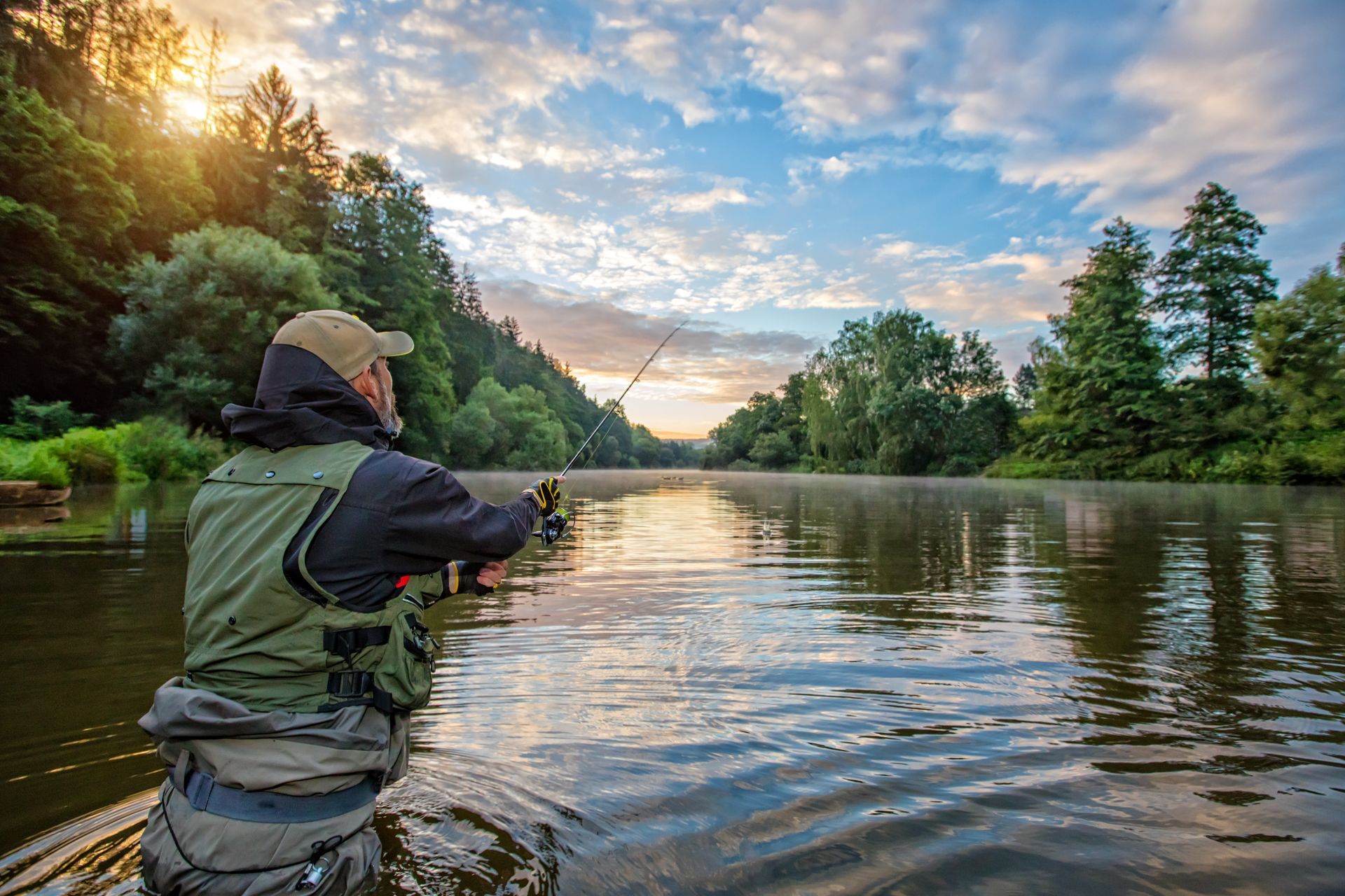 A person in a fishing vest and cap casts a line into a calm, reflective river surrounded by trees at sunrise.