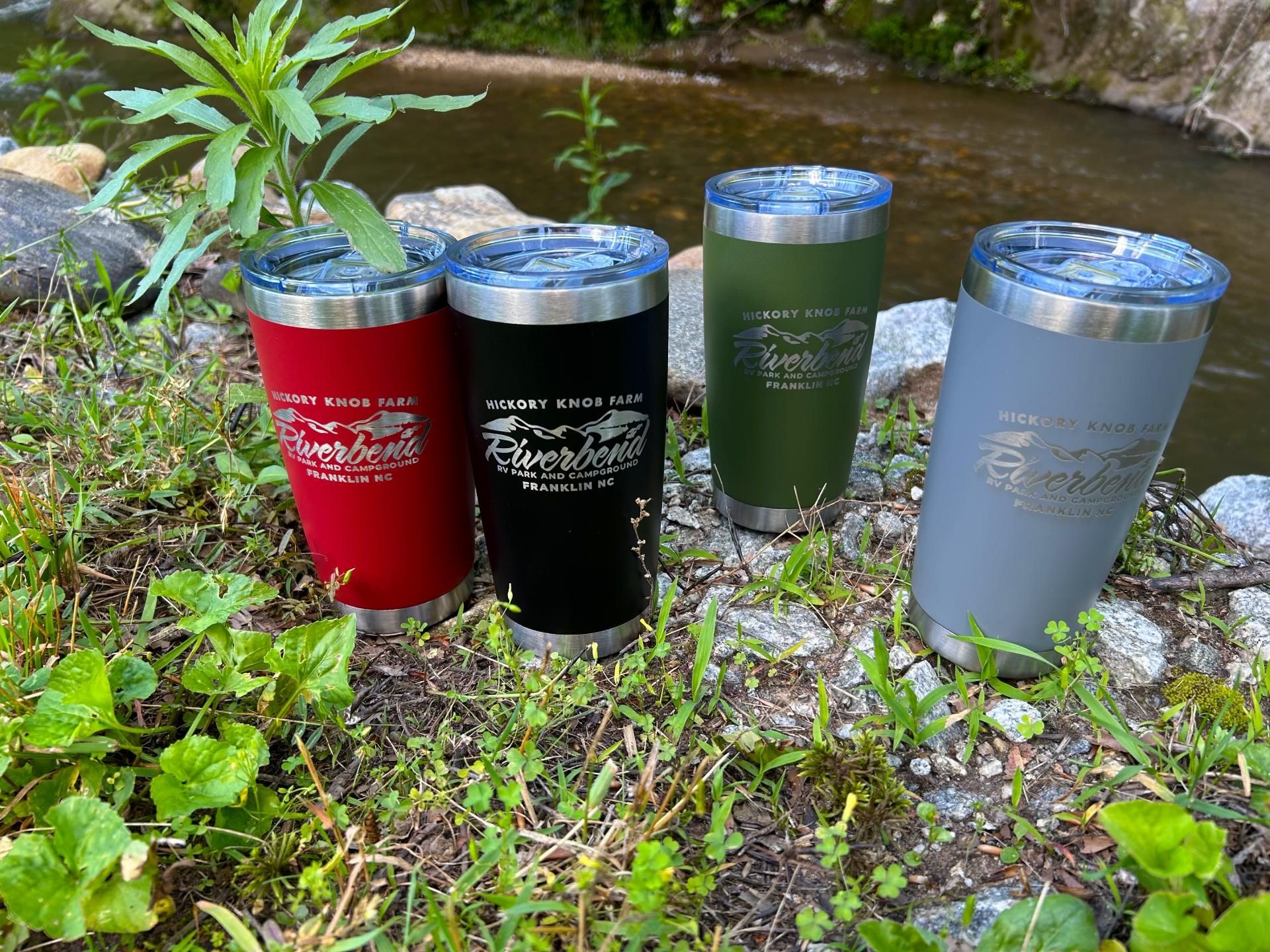 Four insulated tumblers in red, black, olive green, and gray with logos, outdoors near a creek.