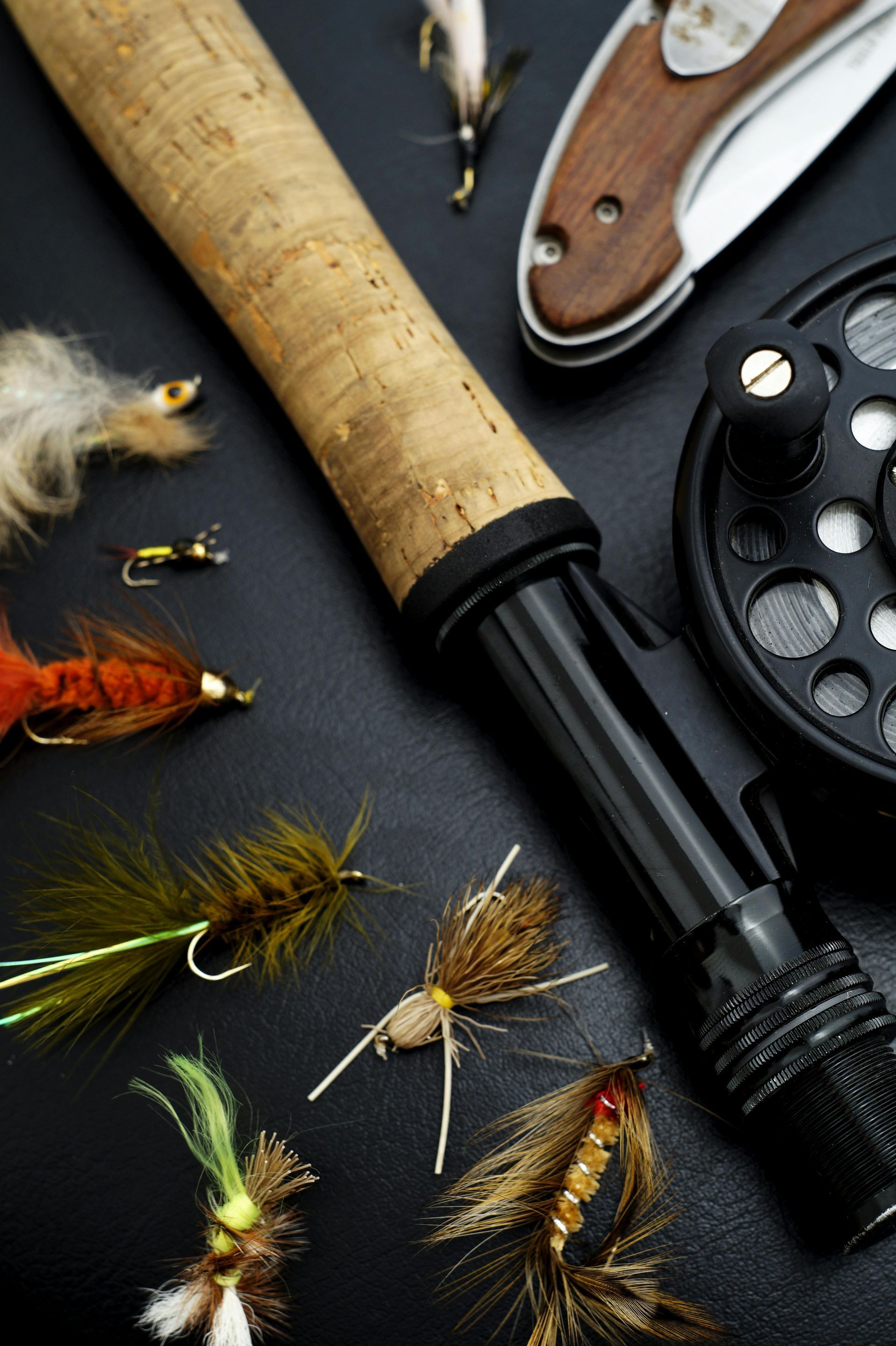 Fly fishing setup on black surface, including rod, reel, flies, and folding knife.