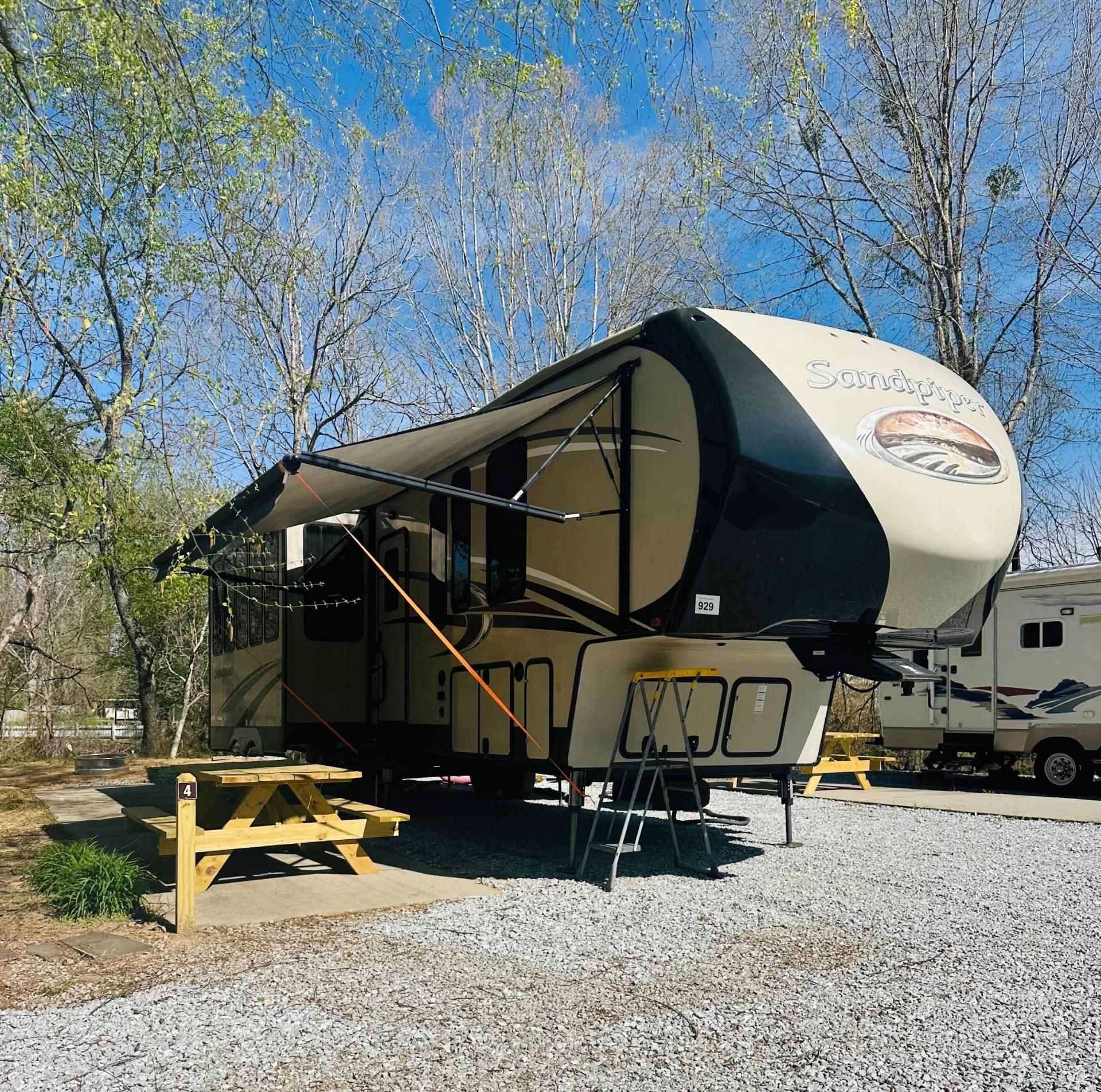 A large fifth-wheel RV with awning extended sits in a gravel campsite next to a picnic table.